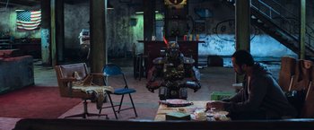 Movie still from “Chappie” (2015), directed by Neill Blomkamp – A robot sitting on top of a chair in a room; Wide shot, High angle