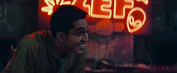 Movie still from “Chappie” (2015), directed by Neill Blomkamp – A young man with glasses is smiling for the camera; Close Up shot, Over the shoulder angle