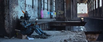 Movie still from “Chappie” (2015), directed by Neill Blomkamp – A person sitting on the ground in front of a building; Wide shot, High angle