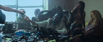 Movie still from “Chappie” (2015), directed by Neill Blomkamp – A man sitting in a chair in front of a bunch of guns; Medium shot, Low angle