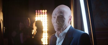 Movie still from “Charlie's Angels” (2019), directed by Elizabeth Banks – A man with a bald head and a suit on in front of lights; Close Up shot, Over the shoulder angle