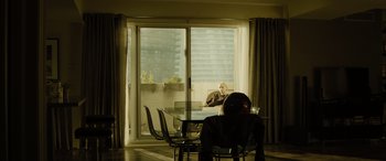 Movie still from “Enemy” (2013), directed by Denis Villeneuve – A person sitting at a table in a room with a sliding glass door; Wide shot, High angle