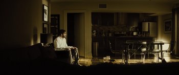 Movie still from “Enemy” (2013), directed by Denis Villeneuve – A man sitting on a chair in a living room; Wide shot, Over the shoulder angle