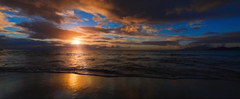 Movie still from “Christmas in Paradise” (2022), directed by Philippe Martinez – The sun is setting over the ocean with a cloudy sky; Extreme Wide shot, High angle