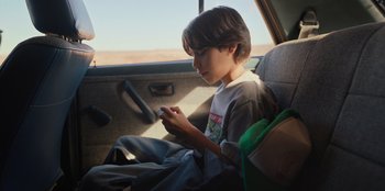 Movie still from “Chupa” (2023), directed by Jonás Cuarón – A boy sitting in the back seat of a car looking at his cell phone; Medium shot, High angle