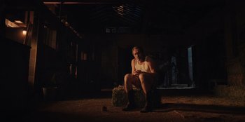 Movie still from “Chupa” (2023), directed by Jonás Cuarón – A man sitting on top of a bale of hay in the dark; Wide shot, Low angle