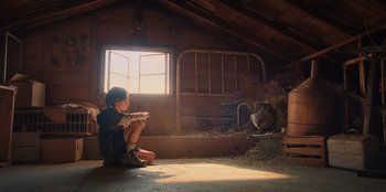Movie still from “Chupa” (2023), directed by Jonás Cuarón – A boy sitting on the ground in a barn looking out a window at an animal; Wide shot, Low angle