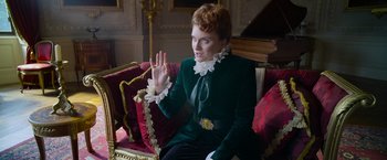Movie still from “Cinderella” (2021), directed by Kay Cannon – A woman sitting on top of a red couch wearing a green jacket; Medium shot, Low angle
