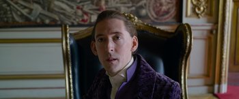 Movie still from “Cinderella” (2021), directed by Kay Cannon – A man in a purple jacket sitting in a chair; Close Up shot, Low angle