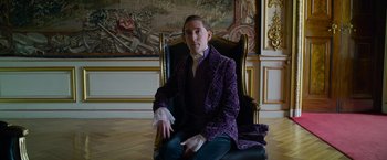 Movie still from “Cinderella” (2021), directed by Kay Cannon – A man sitting in a chair wearing a purple jacket; Medium shot, Low angle