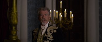 Movie still from “Cinderella” (2021), directed by Kay Cannon – An older man wearing a suit and tie in front of candles; Medium shot, Over the shoulder angle