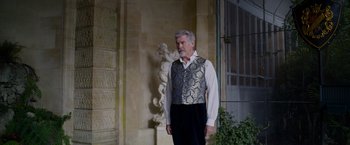 Movie still from “Cinderella” (2021), directed by Kay Cannon – An older man wearing a vest standing in front of a statue; Medium shot, Low angle