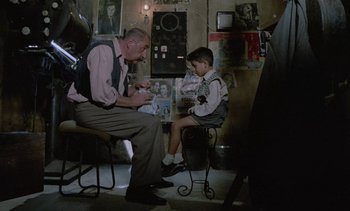 Movie still from “Cinema Paradiso” (1988), directed by Giuseppe Tornatore – An older man sitting next to a young boy in a room; Medium shot, Over the shoulder angle