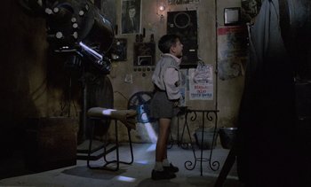 Movie still from “Cinema Paradiso” (1988), directed by Giuseppe Tornatore – A young boy standing in a room next to a chair; Wide shot, High angle