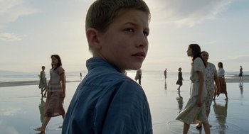 Movie still from “The Tree of Life” (2011), directed by Terrence Malick – A boy standing on the beach with a bunch of people in the background; Close Up shot, Low angle