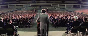 Movie still from “Close Encounters of the Third Kind” (1977), directed by Steven Spielberg – A man is standing at a podium in front of an audience; Extreme Wide shot, High angle