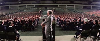 Movie still from “Close Encounters of the Third Kind” (1977), directed by Steven Spielberg – A man standing at a podium in front of an audience; Wide shot, High angle
