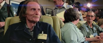 Movie still from “Close Encounters of the Third Kind” (1977), directed by Steven Spielberg – An older man sitting on a seat with a visitor pass on his shoulder; Medium shot, Over the shoulder angle