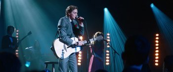 Movie still from “Clouds” (2020), directed by Justin Baldoni – A man in a suit and tie holding a microphone and a woman in a blue dress; Medium shot, Low angle