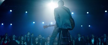 Movie still from “Clouds” (2020), directed by Justin Baldoni – A man sitting on top of a stool holding a guitar in front of an audience; Wide shot, Over the shoulder angle