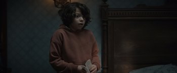 Movie still from “Cobweb” (2023), directed by Samuel Bodin – A young person holding a book in a room; Medium shot, Over the shoulder angle