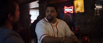 Movie still from “Cocaine Bear” (2023), directed by Elizabeth Banks – A man in a baseball uniform sitting at a table in front of a neon sign; Medium shot, Over the shoulder angle