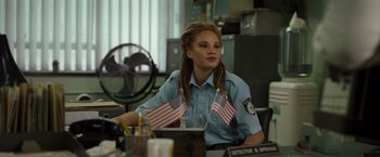 Movie still from “Cocaine Bear” (2023), directed by Elizabeth Banks – A police officer sitting at a desk with two flags; Medium shot, Over the shoulder angle