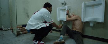 Movie still from “Cocaine Bear” (2023), directed by Elizabeth Banks – A man sitting on the ground next to a urinal; Medium shot, High angle
