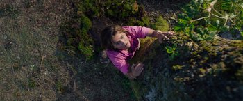 Movie still from “Cocaine Bear” (2023), directed by Elizabeth Banks – A woman in a pink shirt is climbing on a rock; Medium shot, Overhead angle