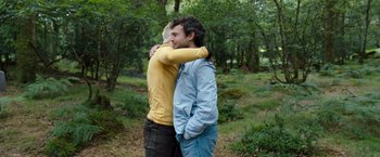 Movie still from “Cocaine Bear” (2023), directed by Elizabeth Banks – Two people hugging in a wooded area with trees in the background; Medium shot, Over the shoulder angle