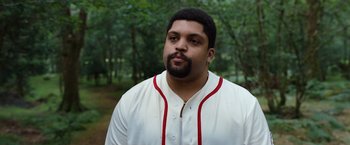 Movie still from “Cocaine Bear” (2023), directed by Elizabeth Banks – A man in a baseball uniform standing in front of a forest; Close Up shot, Low angle