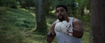 Movie still from “Cocaine Bear” (2023), directed by Elizabeth Banks – A man holding a gun in his hand while standing in the woods; Medium shot, Over the shoulder angle