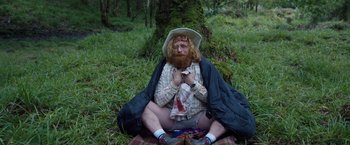 Movie still from “Cocaine Bear” (2023), directed by Elizabeth Banks – A man sitting on the ground in the grass; Wide shot, High angle