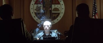 Movie still from “The Usual Suspects” (1995), directed by Bryan Singer – A man sitting at a desk in front of microphones with smoke coming out of his head; Medium shot, Low angle