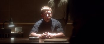 Movie still from “The Usual Suspects” (1995), directed by Bryan Singer – A man sitting at a table with his hands crossed; Medium shot, Over the shoulder angle