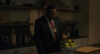 Movie still from “Confess, Fletch” (2022), directed by Greg Mottola – A man standing in a kitchen next to a bowl of fruit; Medium shot, High angle