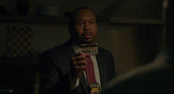 Movie still from “Confess, Fletch” (2022), directed by Greg Mottola – A man in a suit and tie holding a credit card; Close Up shot, Over the shoulder angle