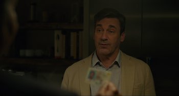 Movie still from “Confess, Fletch” (2022), directed by Greg Mottola – A man in a beige jacket is holding up a card; Close Up shot, Over the shoulder angle