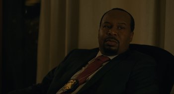 Movie still from “Confess, Fletch” (2022), directed by Greg Mottola – A man wearing a suit and tie sitting in a chair; Close Up shot, Over the shoulder angle