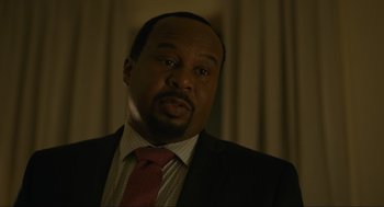 Movie still from “Confess, Fletch” (2022), directed by Greg Mottola – A man wearing a suit and tie in front of a curtain; Close Up shot, Over the shoulder angle