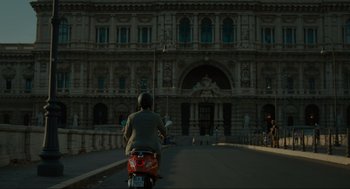 Movie still from “Confess, Fletch” (2022), directed by Greg Mottola – A person riding a motorcycle down a street; Extreme Wide shot, Low angle