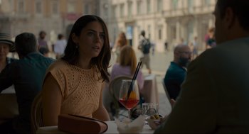 Movie still from “Confess, Fletch” (2022), directed by Greg Mottola – A woman sitting at a table with a glass of wine; Medium shot, Over the shoulder angle