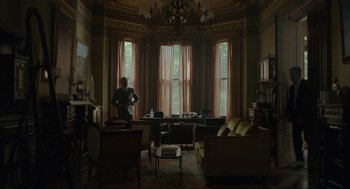 Movie still from “Confess, Fletch” (2022), directed by Greg Mottola – A woman standing in a living room next to a large window; Wide shot, Low angle