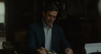 Movie still from “Confess, Fletch” (2022), directed by Greg Mottola – A man sitting at a table with food in front of him; Medium shot, Over the shoulder angle