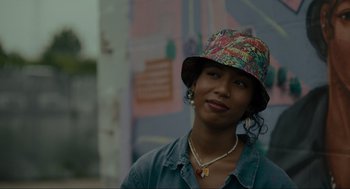 Movie still from “Confess, Fletch” (2022), directed by Greg Mottola – A woman wearing a colorful hat and a necklace; Close Up shot, Over the shoulder angle