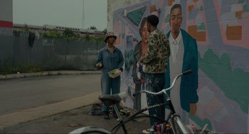 Movie still from “Confess, Fletch” (2022), directed by Greg Mottola – A group of people standing in front of a mural; Wide shot, Over the shoulder angle