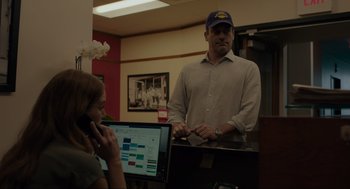 Movie still from “Confess, Fletch” (2022), directed by Greg Mottola – A man standing in front of a laptop computer next to a woman; Medium shot, Over the shoulder angle