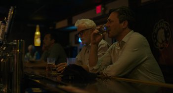 Movie still from “Confess, Fletch” (2022), directed by Greg Mottola – Two men sitting at a bar drinking and smoking; Medium shot, Over the shoulder angle
