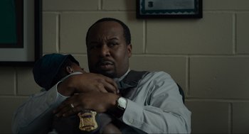 Movie still from “Confess, Fletch” (2022), directed by Greg Mottola – A man in a suit and tie holding a police officer's badge; Close Up shot, High angle