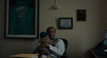 Movie still from “Confess, Fletch” (2022), directed by Greg Mottola – A man sitting in a chair holding a baby; Medium shot, Over the shoulder angle